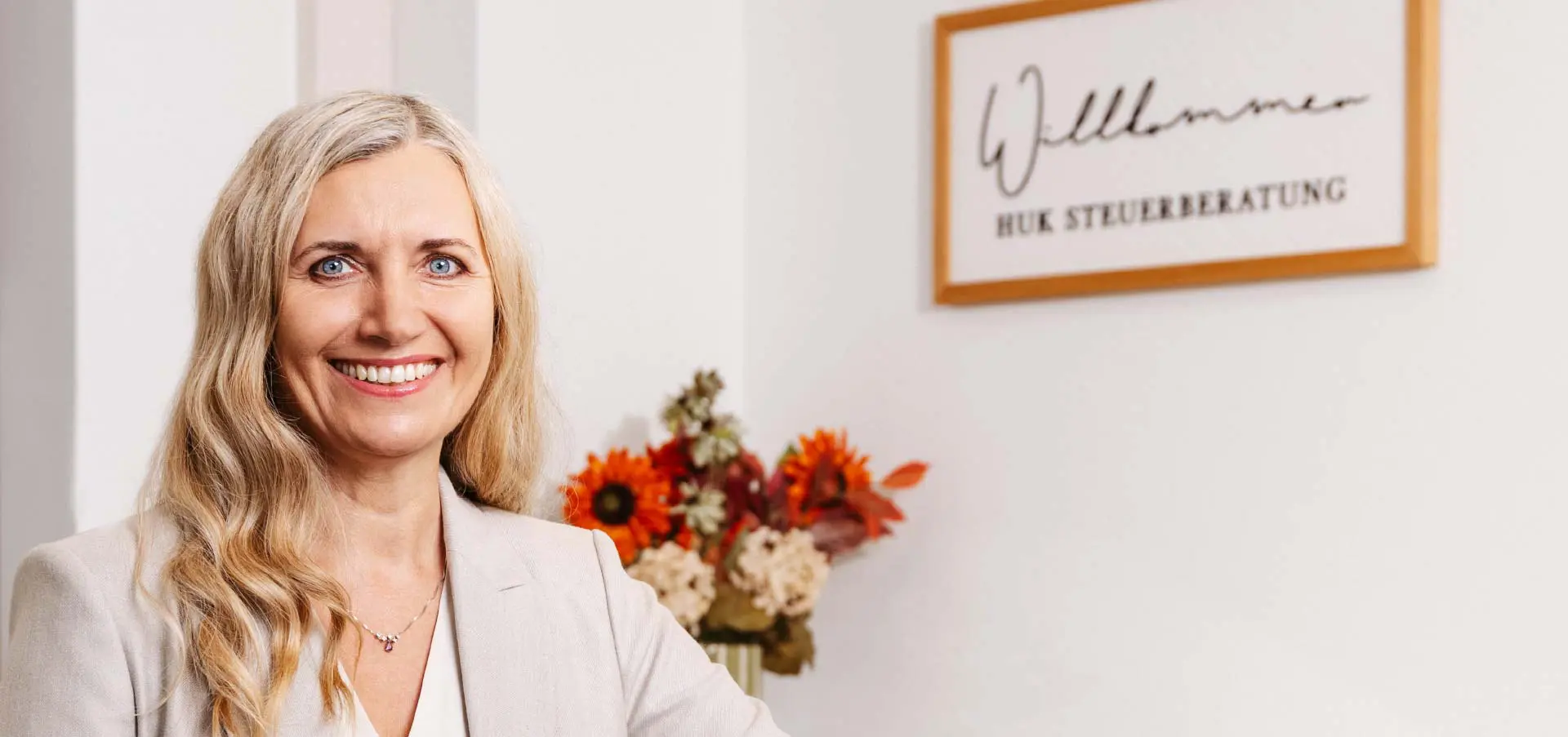 Katharina Huk, Steuerberaterin, schaut lächelnd in die Kamera, neben ihr ein Blumenstrauss und ein Willkommen-Schild an der Wand.